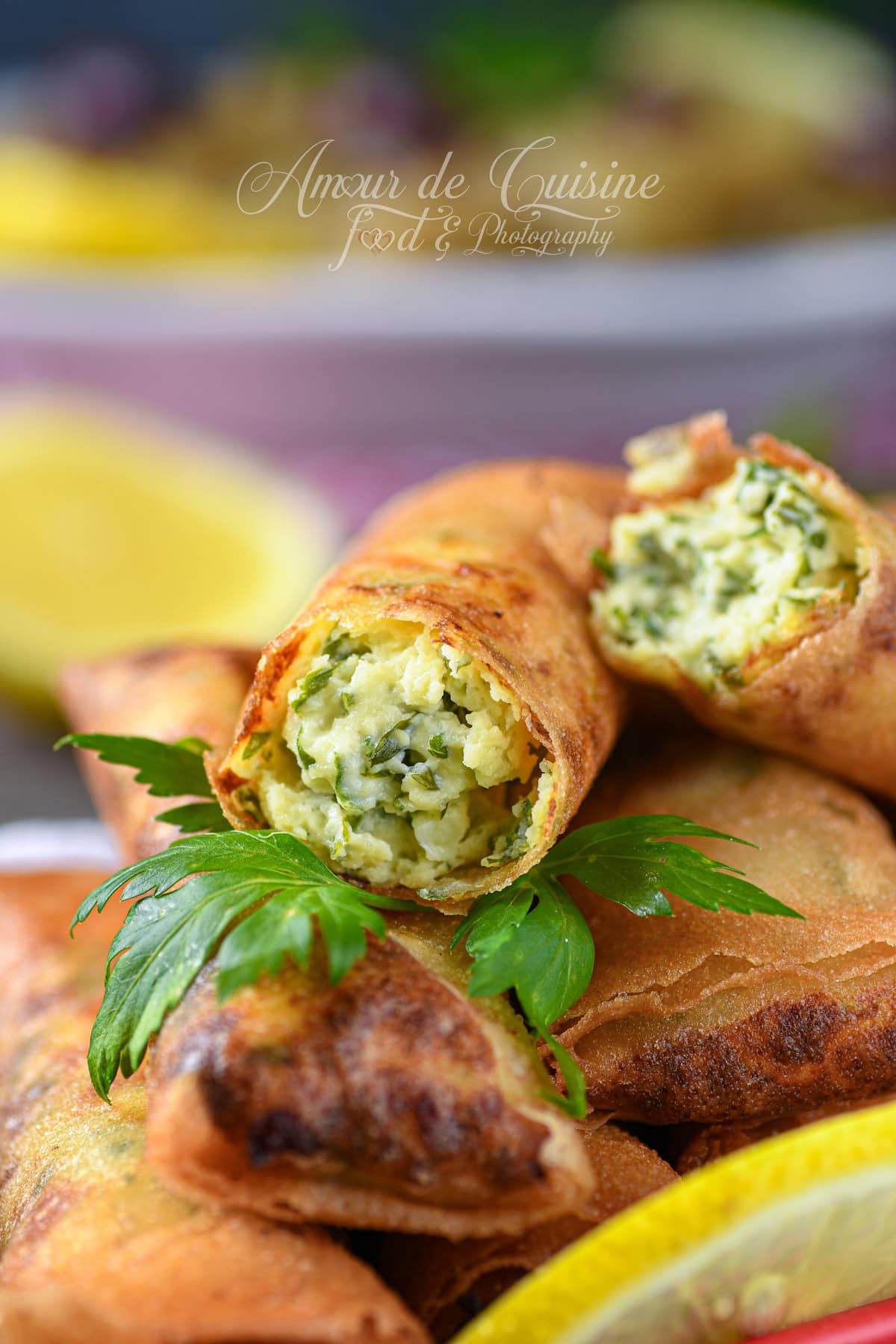 Close-up of golden cheese cigars, with one cut open to show a creamy cheese-and-parsley filling, stacked on other boureks with parsley leaves and lemon in the background.