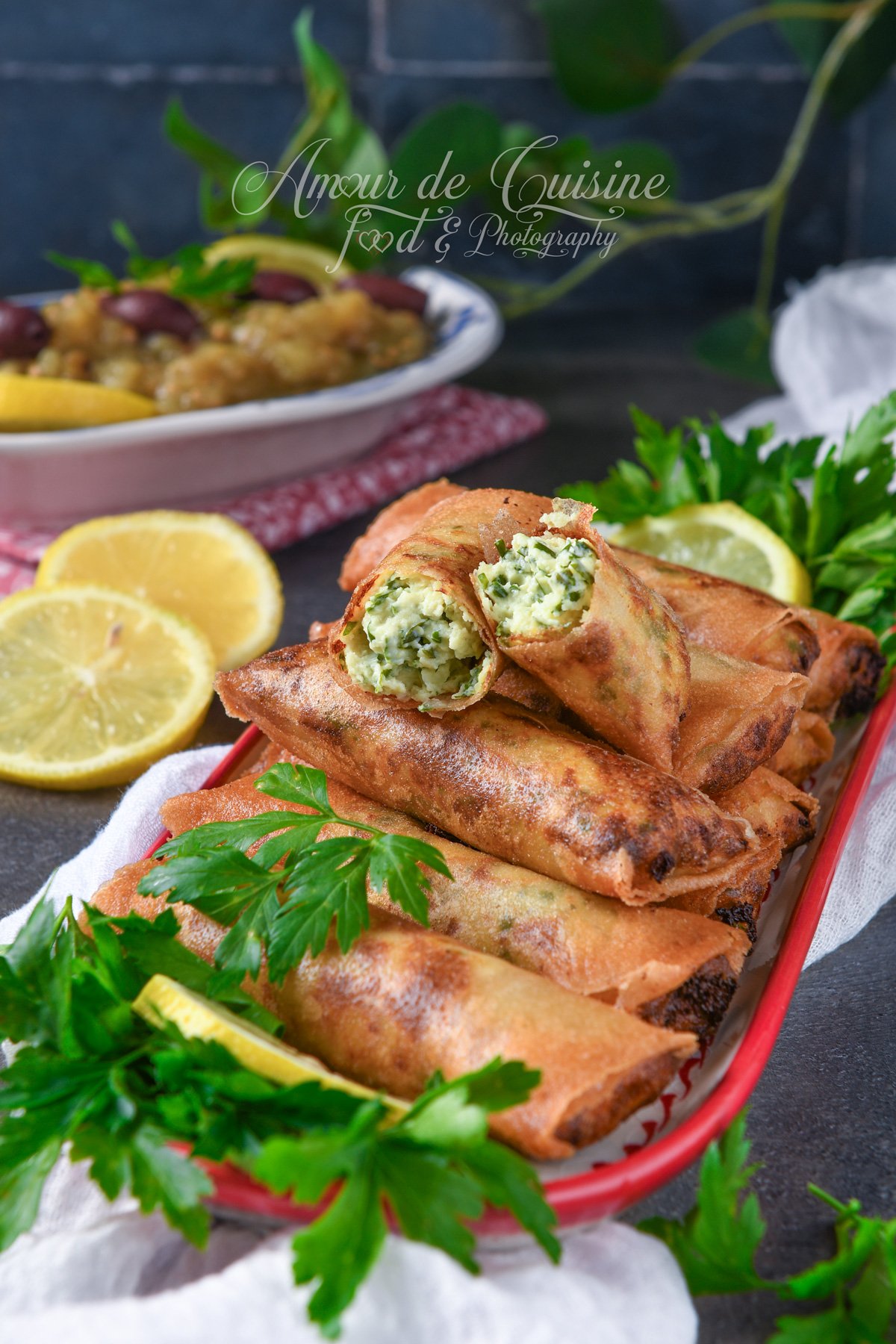 Golden, crispy cheese cigars on a serving platter with fresh parsley and lemon wedges, with one cut open to reveal a creamy herb-and-cheese filling.