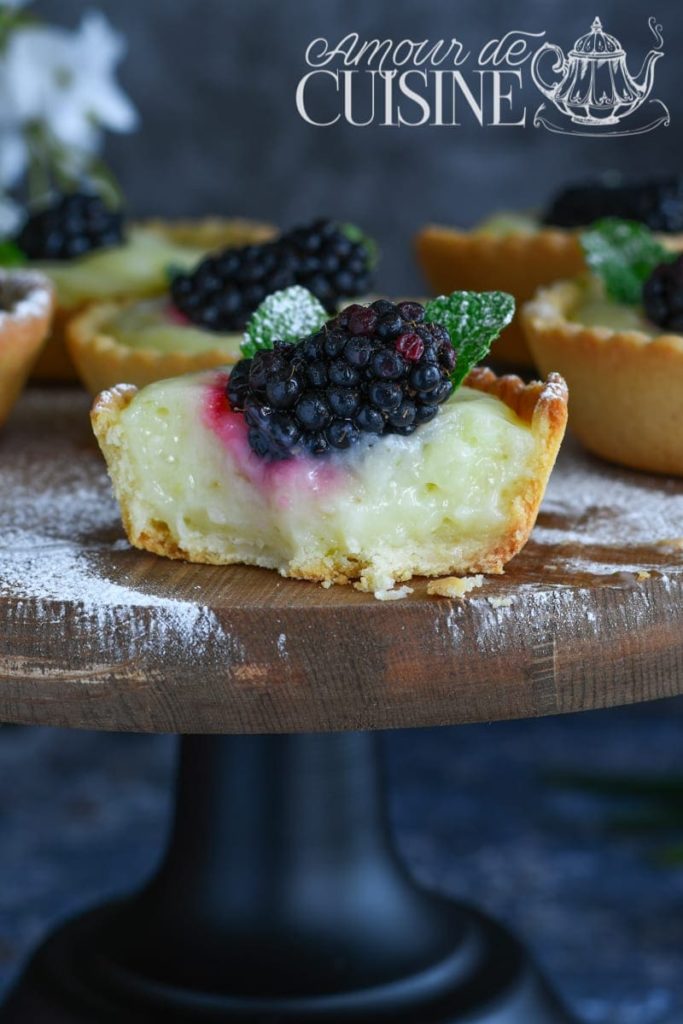 Half-cut mini lemon curd tartlet with creamy filling, topped with a fresh blackberry and mint leaf, displayed on a wooden cake stand dusted with icing sugar.
