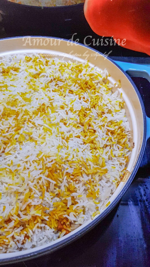 A close-up of a pan filled with layered basmati rice for chicken biryani, featuring vibrant saffron-colored strands and white grains, just before the final cooking stage. The rice is neatly spread in a round, enameled cast iron pot on a stovetop