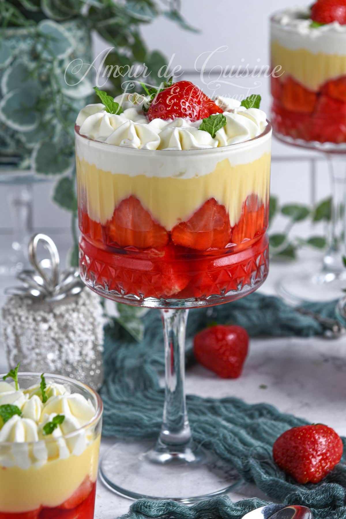 Elegant English trifle dessert with layers of strawberry jelly, vanilla custard, and whipped cream, topped with a fresh strawberry and mint leaves, served in a crystal glass on a dessert table.