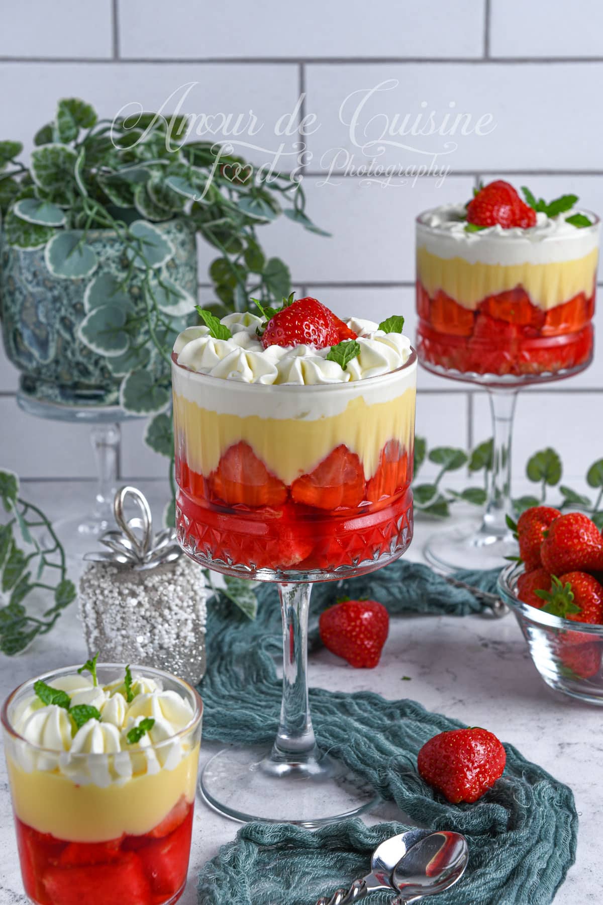 English strawberry trifles in elegant glass cups, layered with red jelly, vanilla custard, and whipped cream, topped with fresh strawberries and mint leaves, displayed on a decorated dessert table.