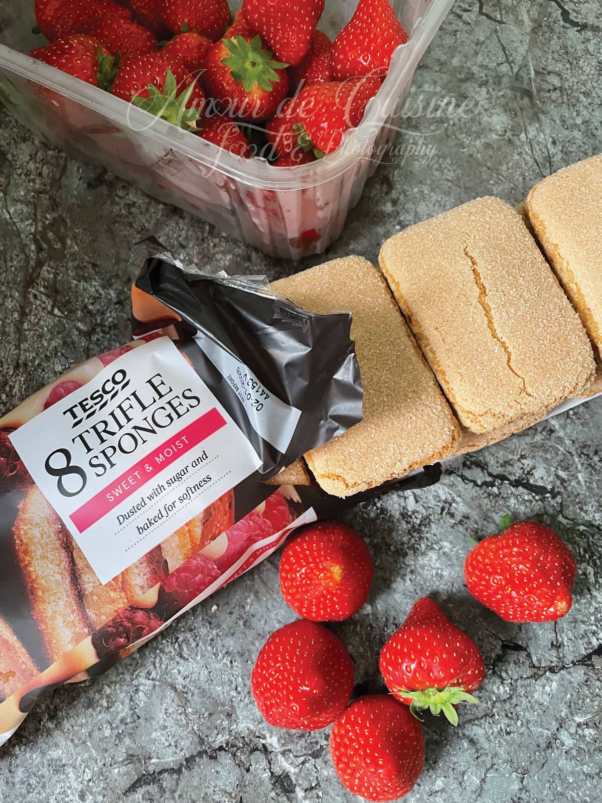 Ingredients for strawberry trifle: fresh strawberries in a container and packaged trifle sponge cakes from Tesco, arranged on a marble countertop.