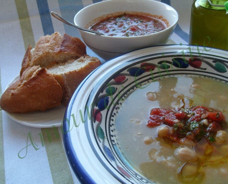 Assiette de doubara de Biskra aux pois chiches, garnie d’une sauce tomate épicée, servie avec du pain, un bol de sauce et une bouteille d’huile d’olive.