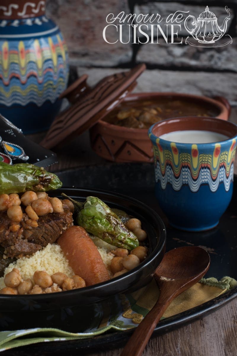 Couscous à l'agneau et aux légumes Assiette de couscous à l’agneau et aux légumes traditionnels, servie avec pois chiches, carotte et poivron vert, accompagnée d’un verre de lait fermenté et d’un tajine en arrière-plan, recette maghrébine authentique conviviale.