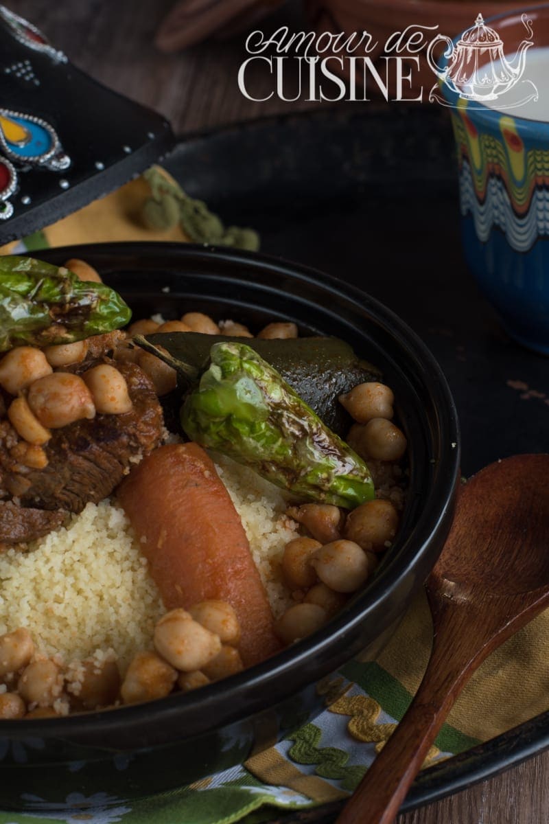 Plat traditionnel de couscous algérien à l’agneau et aux légumes, garni de pois chiches, carotte, courgette et poivron, servi dans un grand plat noir avec semoule légère et sauce parfumée, recette authentique de la cuisine maghrébine.