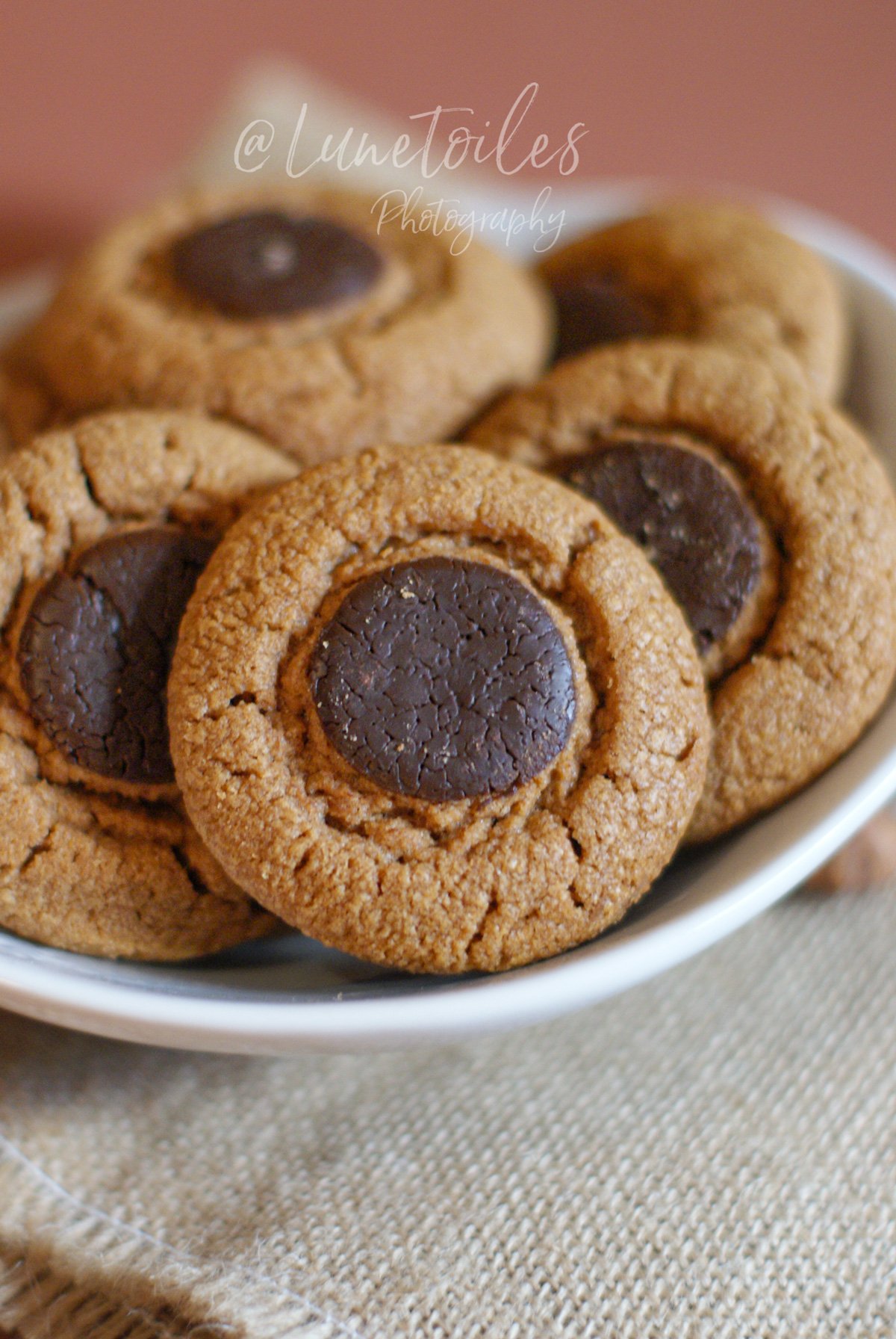 Golden, lightly crackled praline cookies topped with a dark chocolate disc, served in a white bowl on a linen cloth.