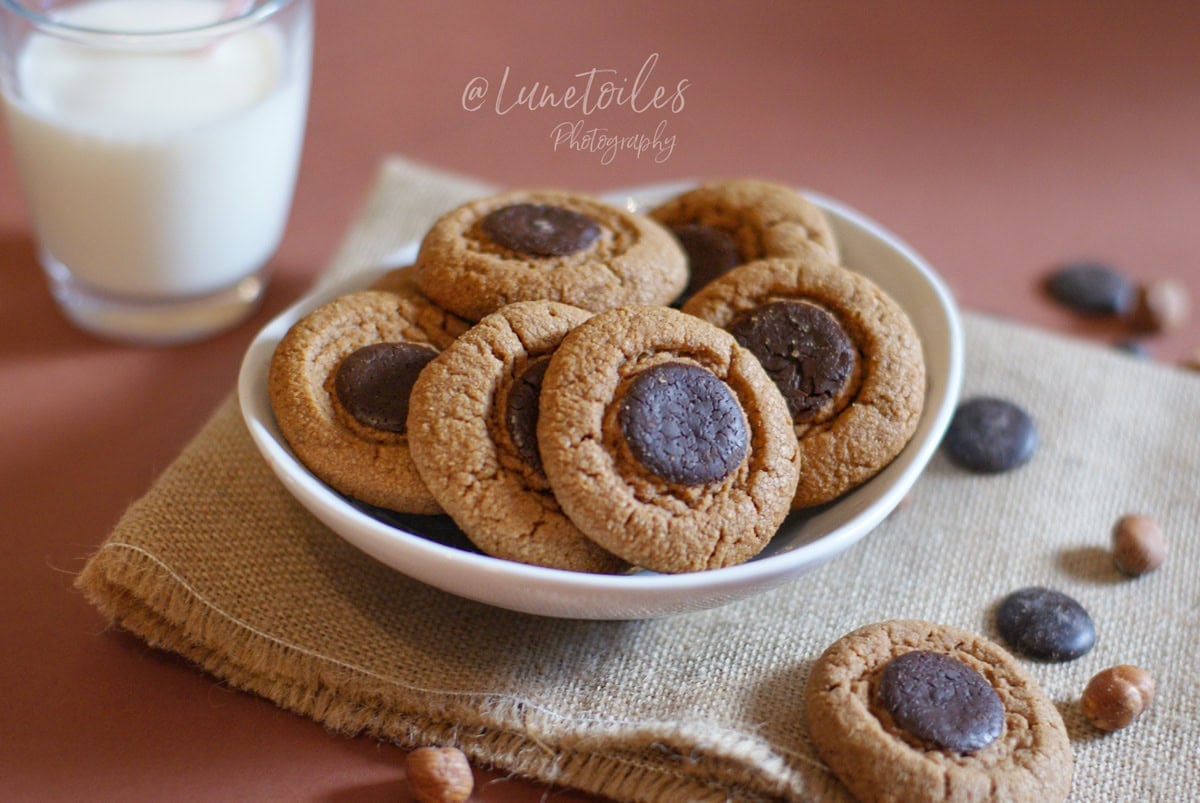 Small bowl of golden, lightly crackled chocolate-praline cookies topped with a dark chocolate disc, set on burlap with a few hazelnuts, with a glass of milk nearby.