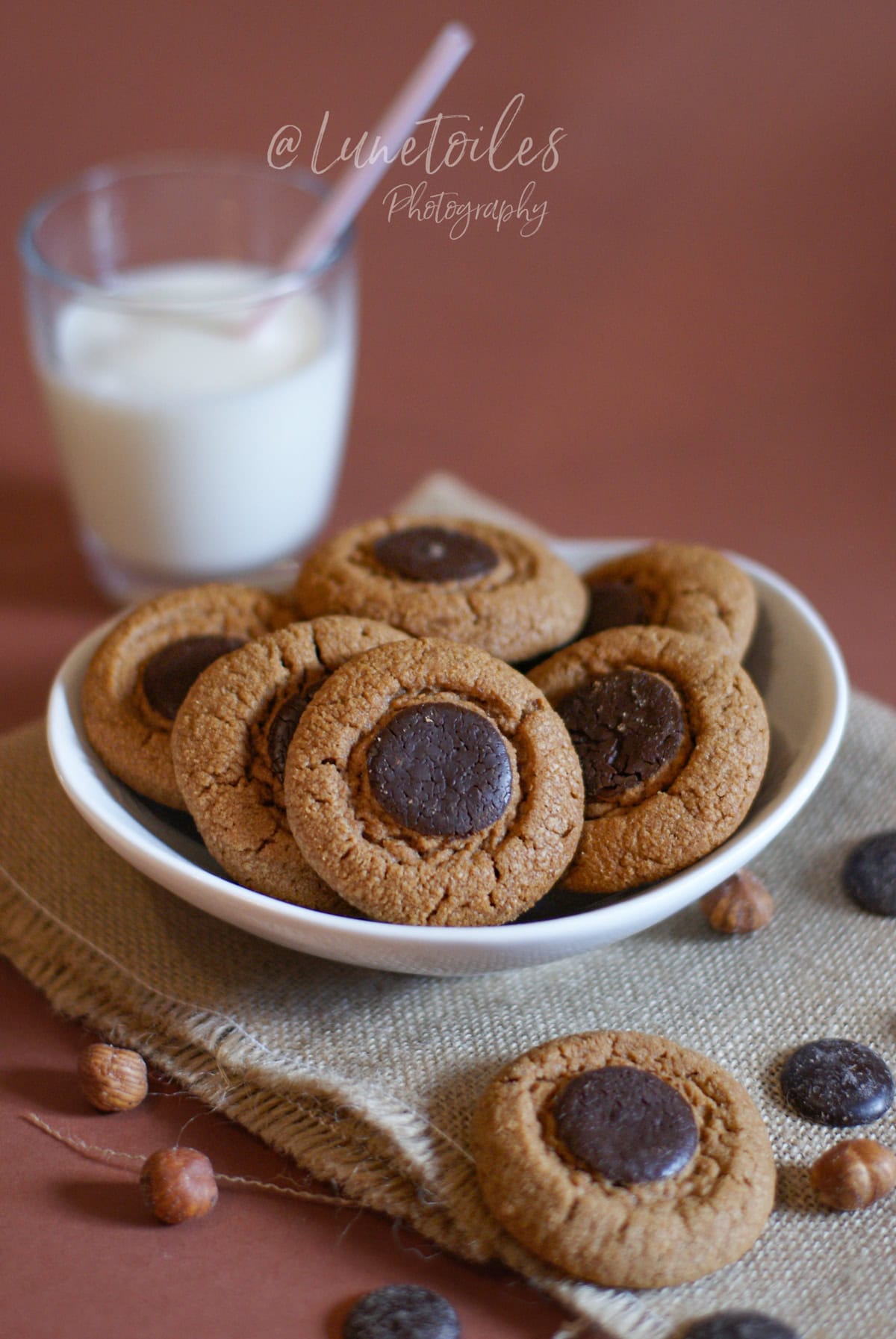 Bowl of golden, lightly crackled chocolate-praline cookies topped with a dark chocolate disc, set on a linen cloth with a few hazelnuts, with a glass of milk in the background.