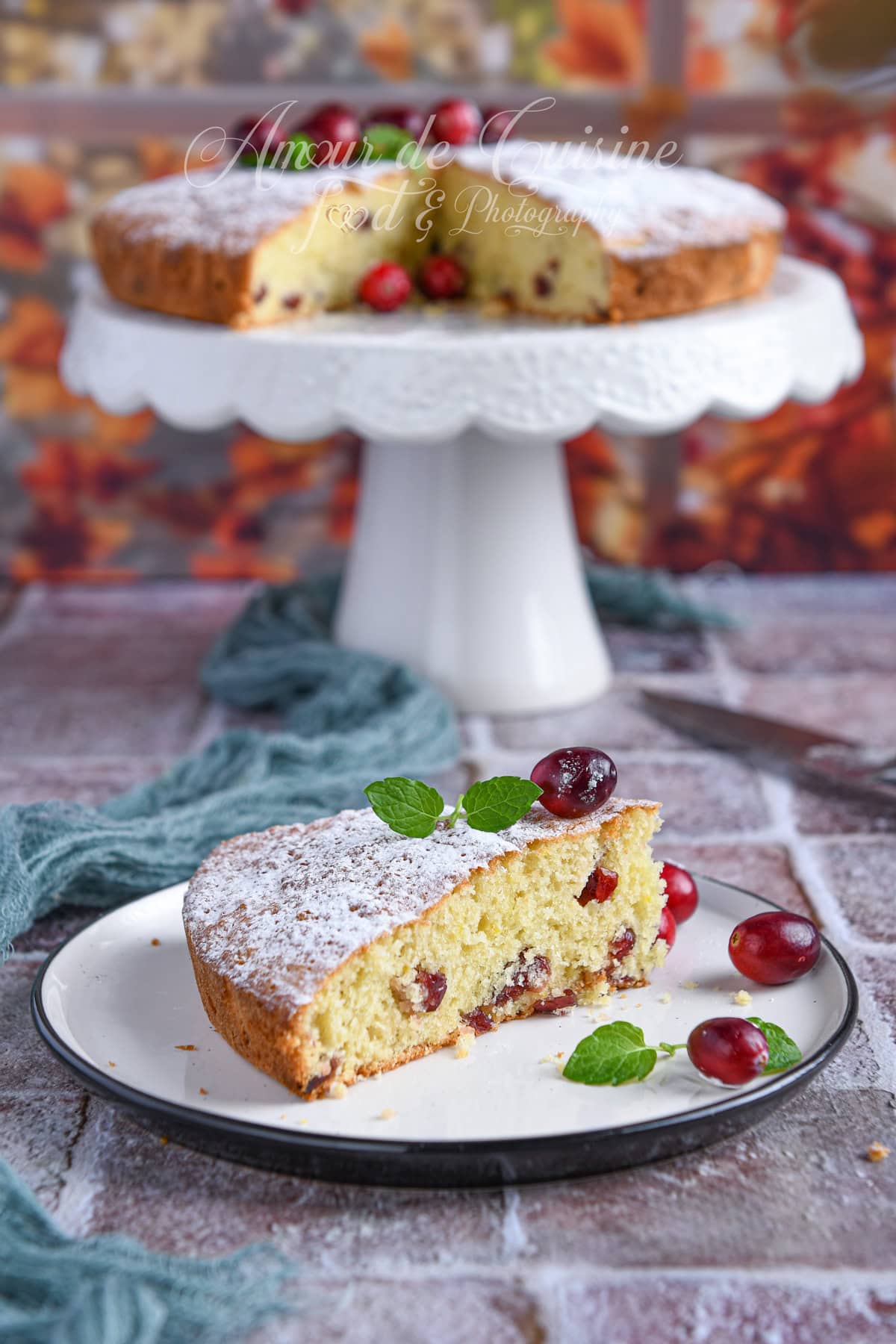 Slice of cranberry cake on a plate with fresh cranberries and mint, with the remaining cake displayed on a white stand in the background against an autumn-themed setting.