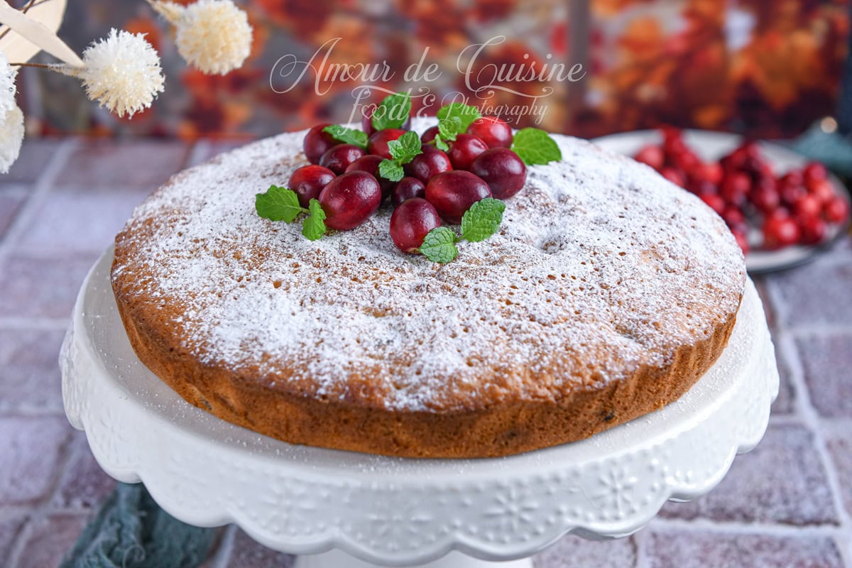 Whole cranberry cake dusted with powdered sugar and topped with fresh cranberries and mint leaves, displayed on a white decorative cake stand with an autumn-colored background.