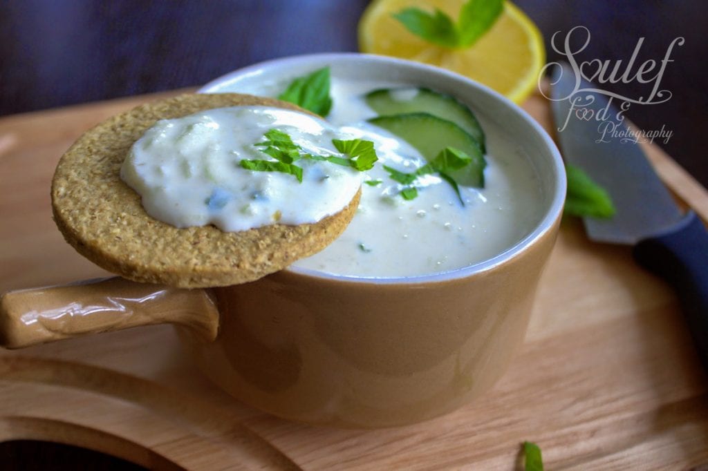 A bowl of creamy cucumber raita garnished with cucumber slices and mint, served with a round cracker dipped in the raita. A lemon half and a knife are visible in the background on a wooden tray