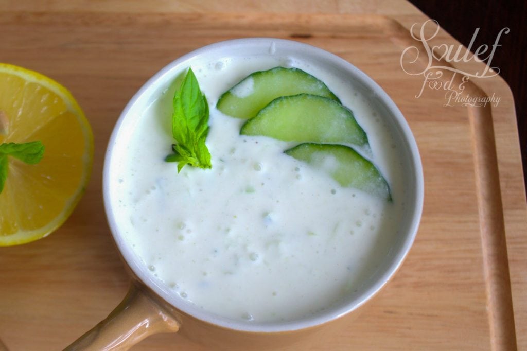 A bowl of creamy cucumber raita garnished with fresh mint leaves and thin cucumber slices, placed on a wooden tray next to a halved lemon.