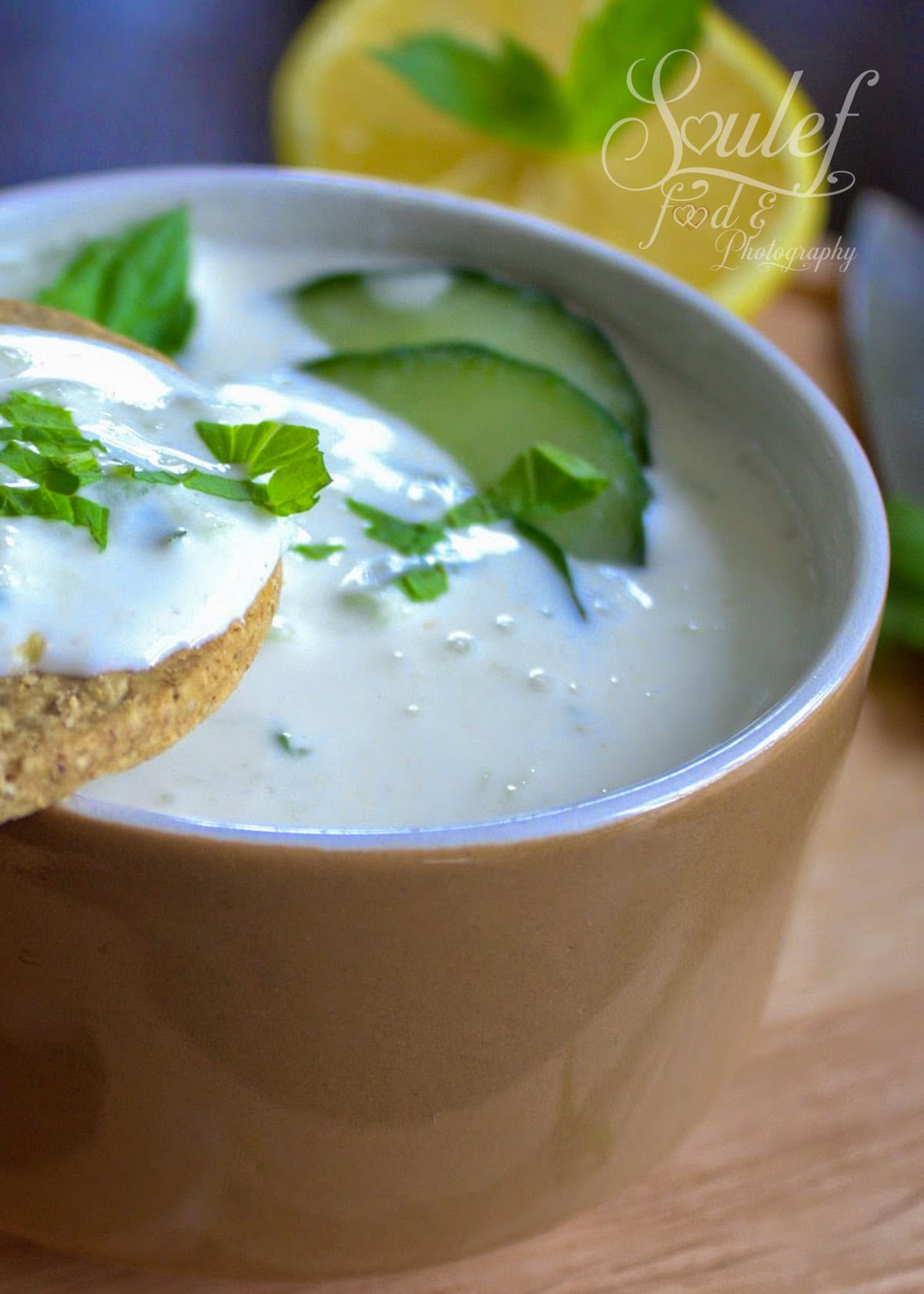 Bol de raita au concombre crémeux, garni de tranches de concombre frais, de feuilles de menthe et accompagné d’un biscuit trempé dans la préparation. Un demi-citron est visible en arrière-plan sur une planche en bois.