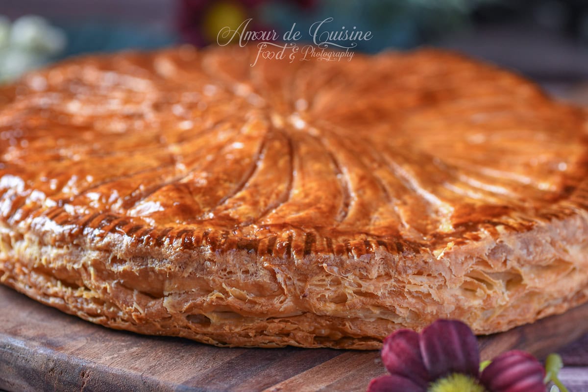 Golden, glossy galette made with express puff pastry, with a knife-scored pattern on top and visible flaky layers along the edge, placed on a wooden board.