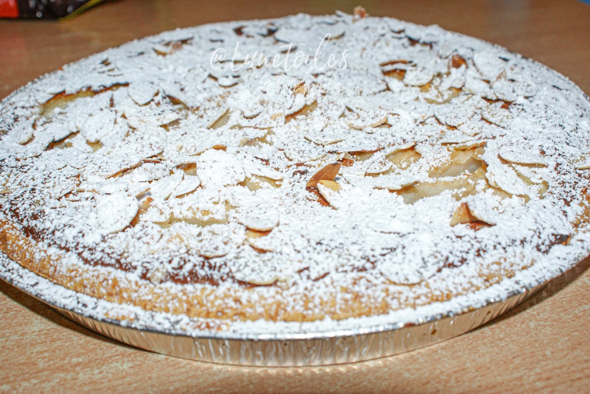 Close-up of a slice of pear amandine tart, generously filled, moist, and topped with powdered sugar and flaked almonds, served on a colorful plate