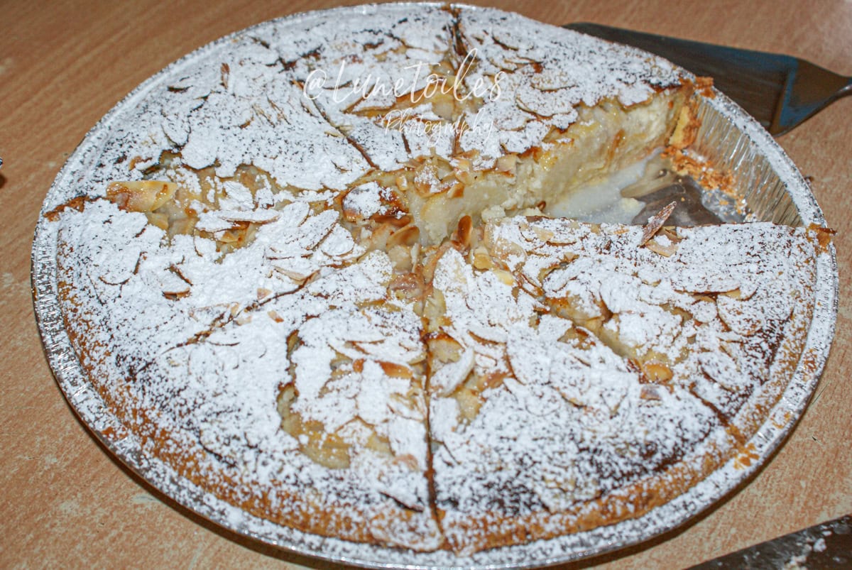 Partially sliced pear amandine tart, cut into generous portions, topped with flaked almonds and dusted with powdered sugar, in an aluminum pan on a countertop.