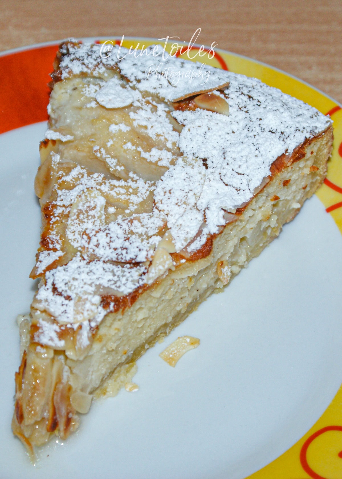 Slice of pear amandine tart topped with flaked almonds and dusted with powdered sugar, served on a colorful plate