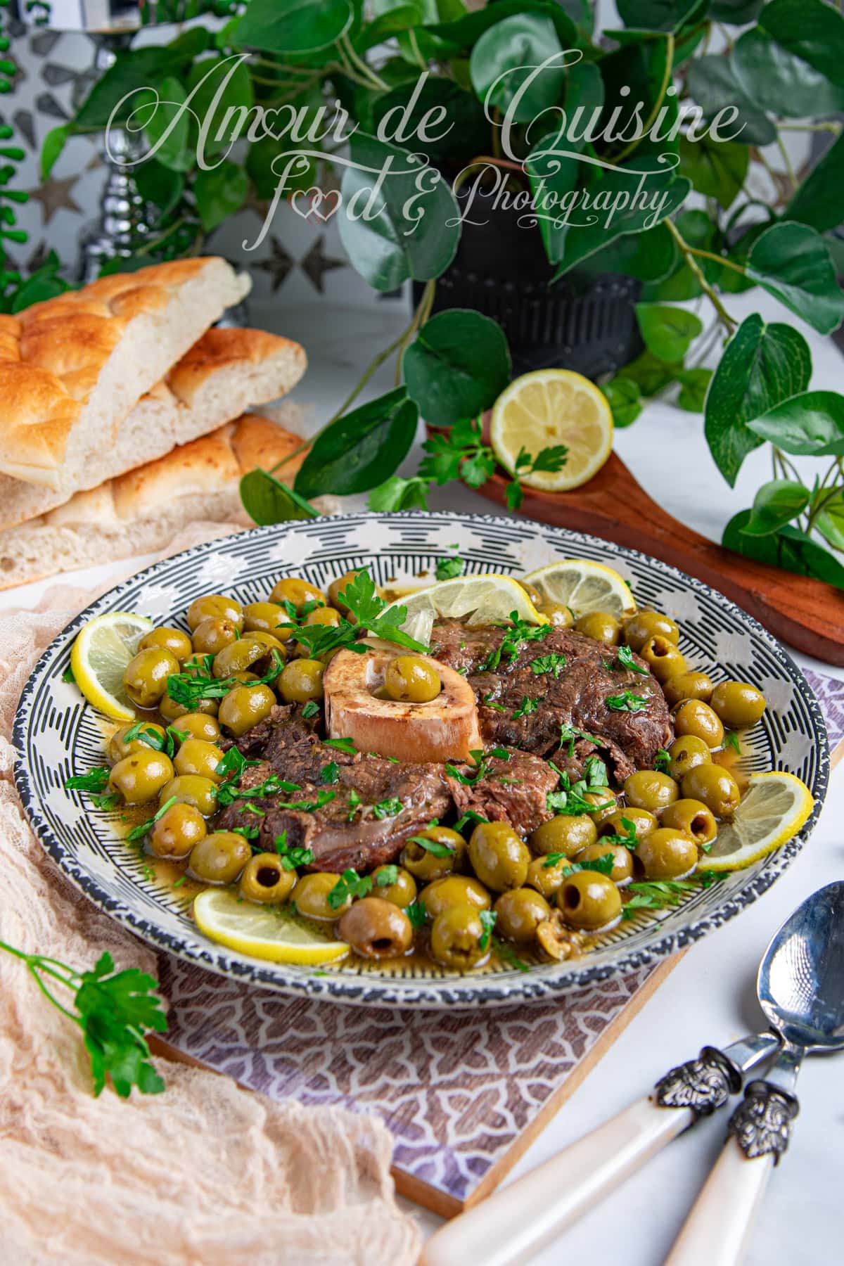 Plat marocain de jarret de bœuf mijoté aux olives vertes et citron confit, garni de persil frais et servi dans une assiette traditionnelle, accompagné de pain maison et décoré de rondelles de citron, sur une table garnie de verdure