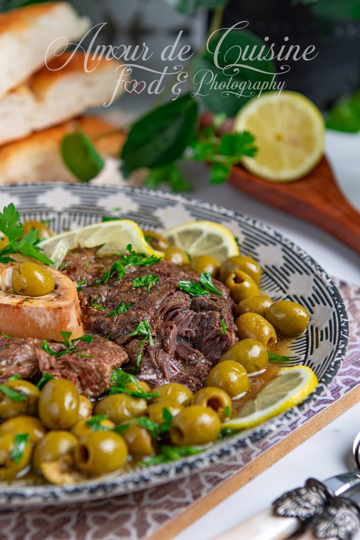 Plat marocain traditionnel de viande de jarret de boeuf mijotée avec des olives vertes, des tranches de citron, et un os à moelle, servi dans une assiette décorative, accompagné de pain frais en arrière-plan