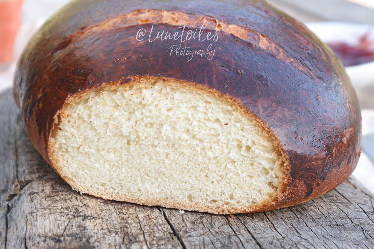 Tourton nantais (pain sucré de Nantes) à la croûte bien dorée et brillante, coupé pour montrer une mie tendre et aérée, posé sur une planche en bois rustique avec un bol de confiture flou en arrière-plan.