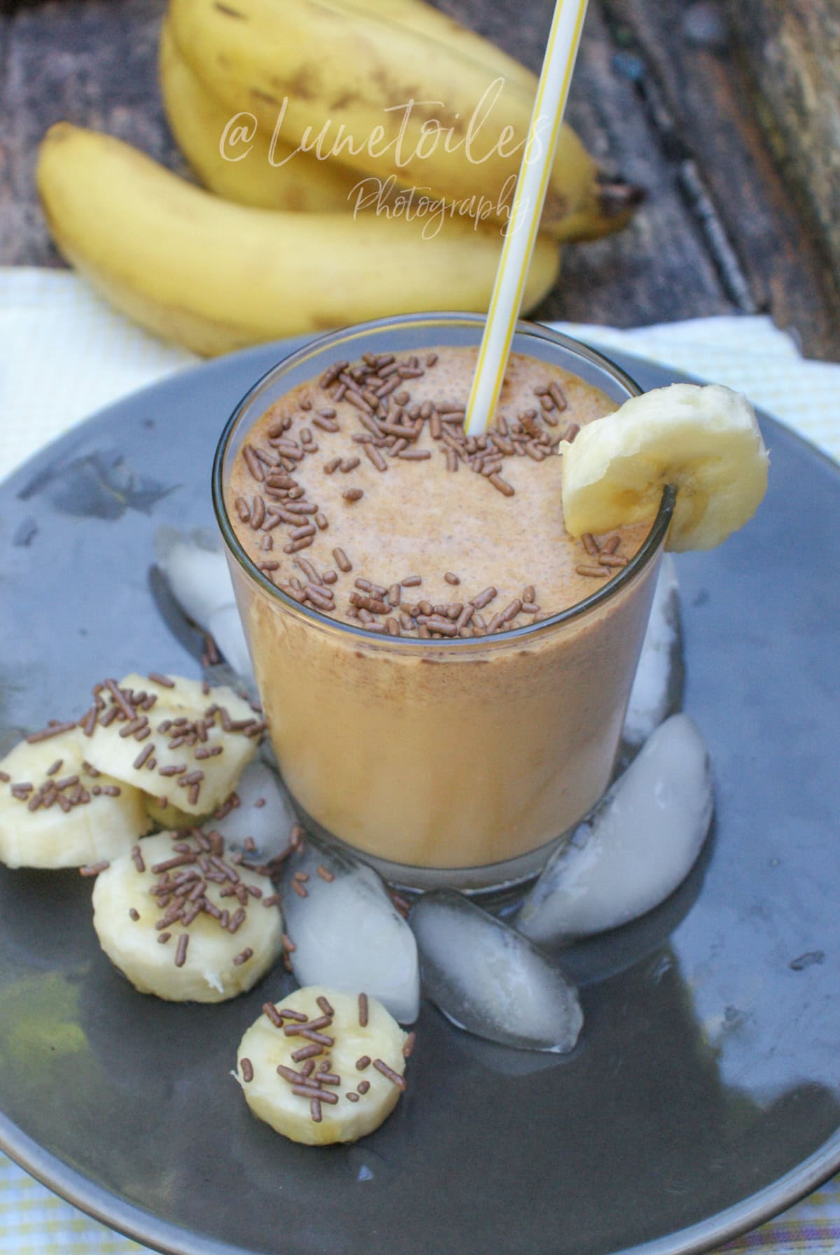 Creamy banana and peanut butter smoothie in a glass, topped with chocolate sprinkles, with a straw and a banana slice garnish, set on a plate with ice cubes and banana slices, whole bananas in the background.