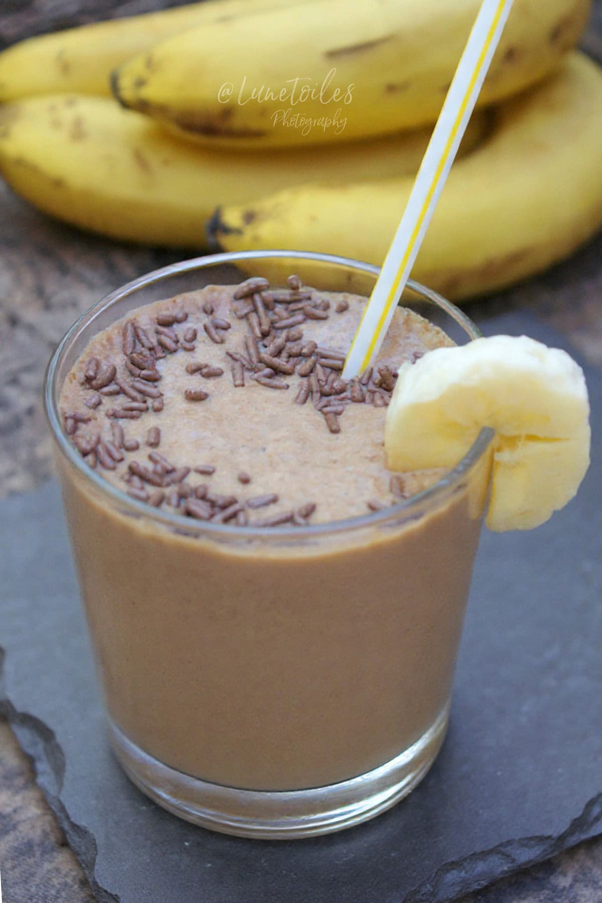 Creamy banana and peanut butter smoothie in a glass, topped with chocolate sprinkles, with a straw and a banana slice on the rim, set on a slate board with bananas in the background.