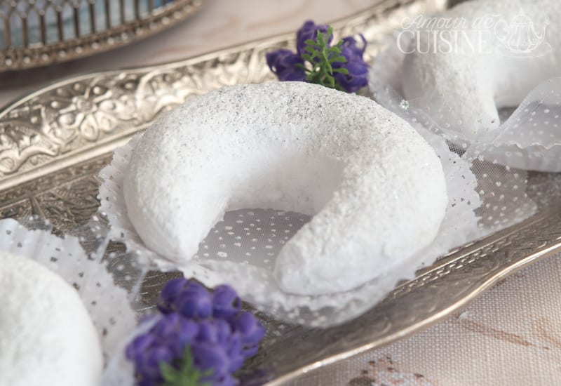 Algerian tcharek msaker crescent cookie coated in powdered sugar, served on an ornate silver tray with a lace doily and purple flowers.