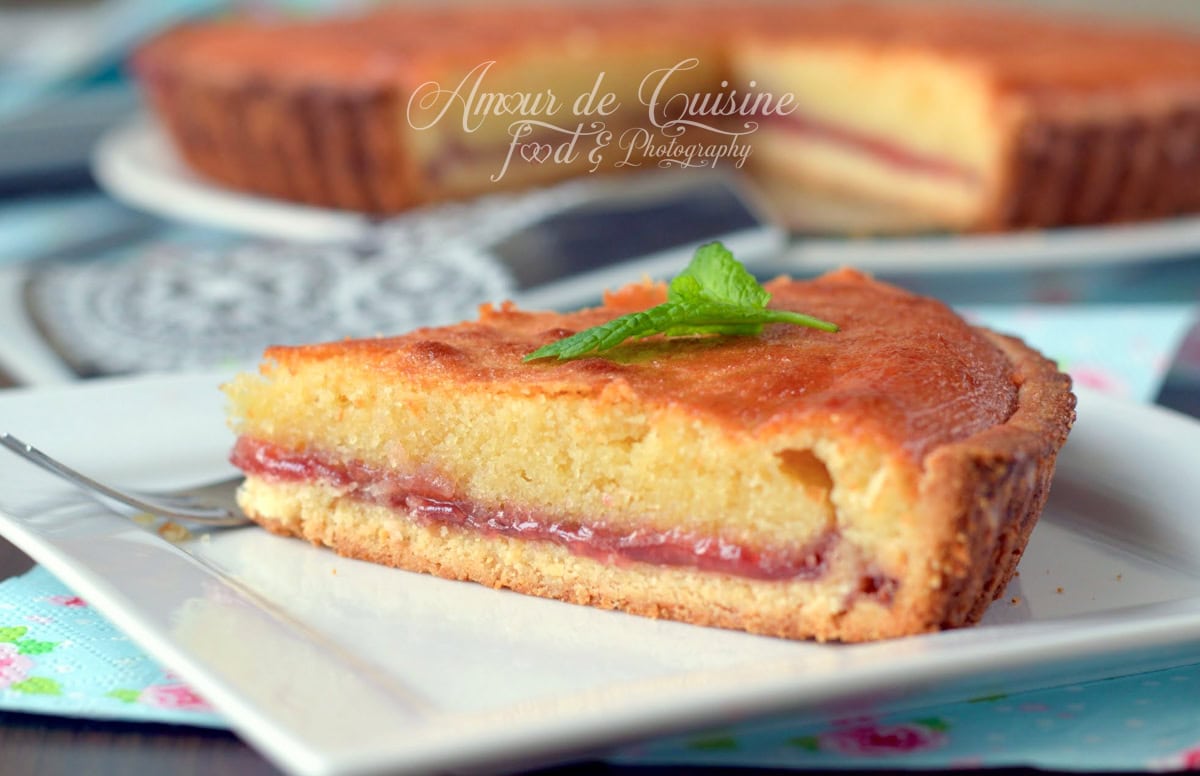 Slice of golden Bakewell tart topped with a layer of raspberry jam and soft almond cream, decorated with a mint leaf on a white plate.