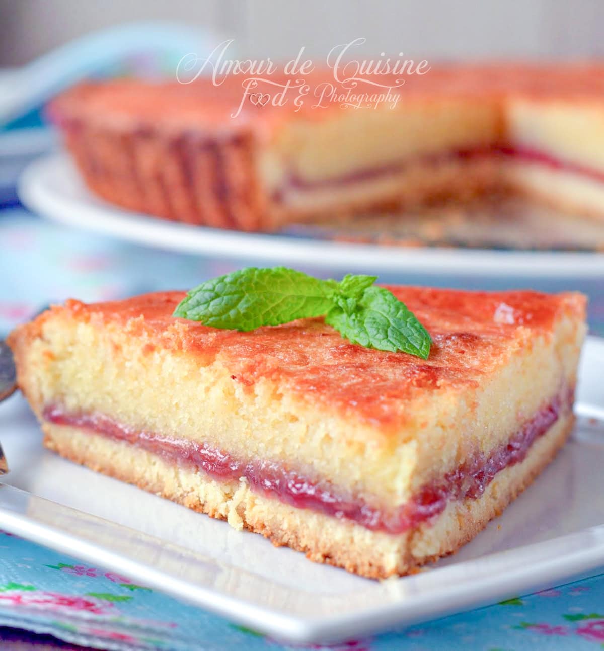 Slice of golden Bakewell tart topped with a layer of red jam and soft almond frangipane, decorated with a fresh mint leaf on a white plate