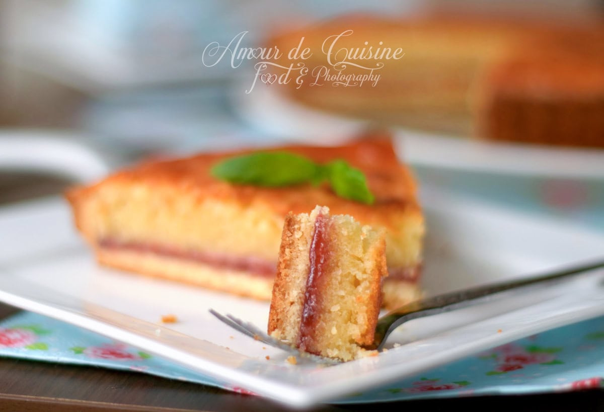 Close-up of a soft Bakewell tart slice with a glossy jam layer and golden frangipane, a piece resting on a fork on a white plate.