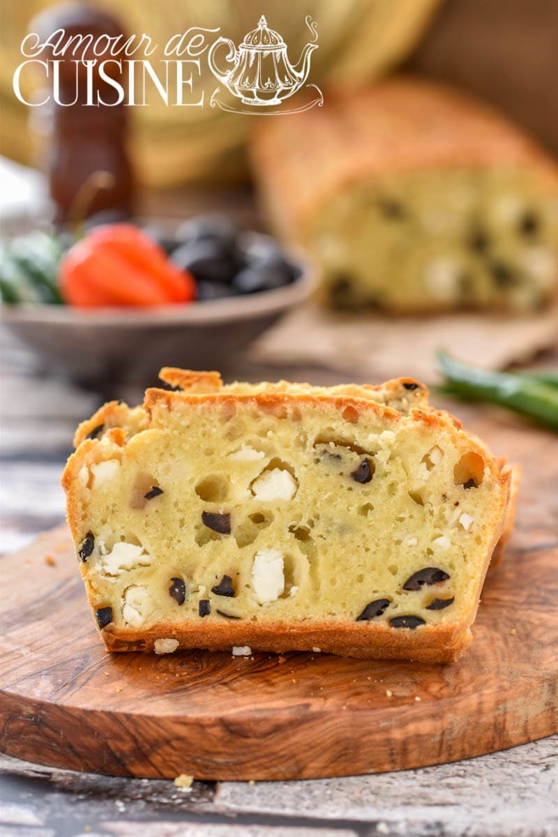 Tranche de cake salé moelleux aux olives noires et à la feta, posée sur une planche en bois. En arrière-plan, un cake entier flou, ainsi qu'un bol d'olives, un piment et des ingrédients méditerranéens