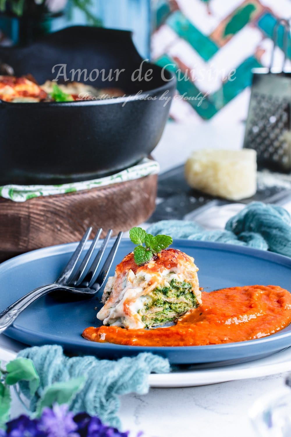 Bouchée de gratin de crêpes à la florentine servie dans une assiette bleue, posée sur un lit de coulis de tomate, avec farce épinards-ricotta visible et une petite feuille de menthe, sur fond de poêle en fonte.