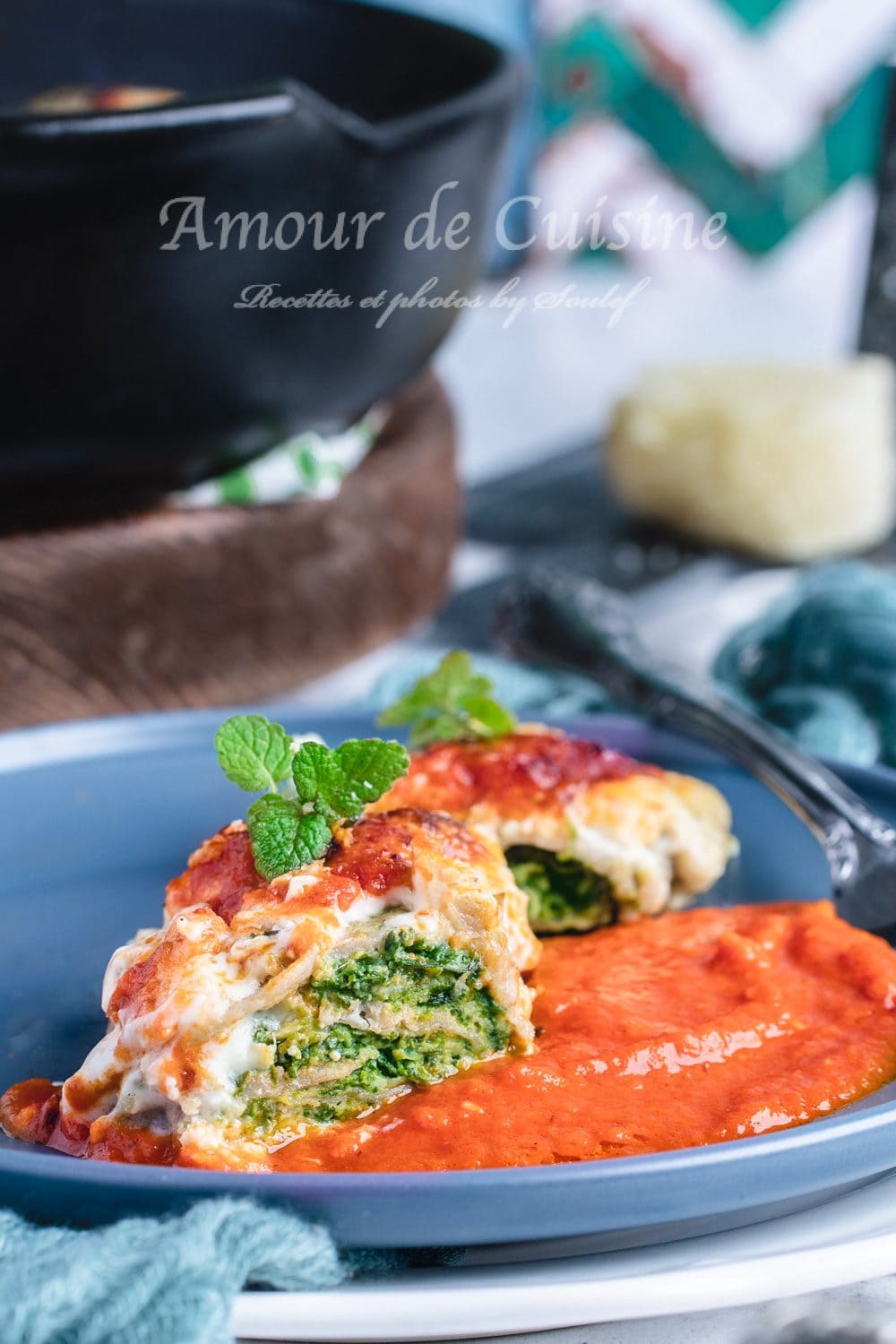 Part de gratin de crêpes à la florentine servie dans une assiette bleue, avec une farce ricotta-épinards visible à la coupe, nappée de béchamel et posée sur un lit de coulis de tomate, décorée de menthe.