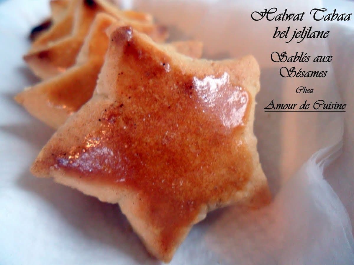 Gâteau sec algérien Halwat Tabaa en forme d’étoile, doré et brillant, présenté en file sur une assiette. Une mention en texte indique : "Halwat Tabaa bel jejlane - Sablés aux Sésames - chez Amour de Cuisine" 