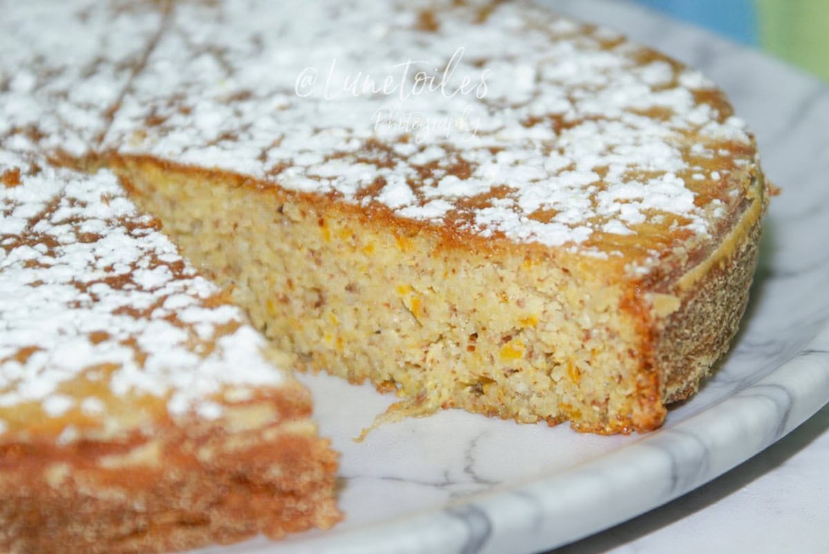 Close-up of a slice of gluten-free orange almond cake, moist and slightly grainy, with a golden top dusted with icing sugar on a white plate.