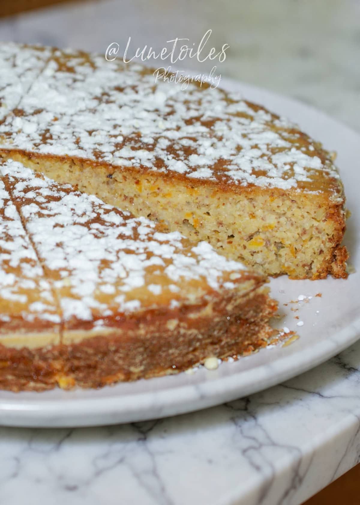 Gluten-free orange cake, moist and golden, dusted with icing sugar. One slice is cut out, revealing a tender crumb with bits of almond. Served on a wicker tray, this homemade dessert is perfect for a comforting sweet treat.