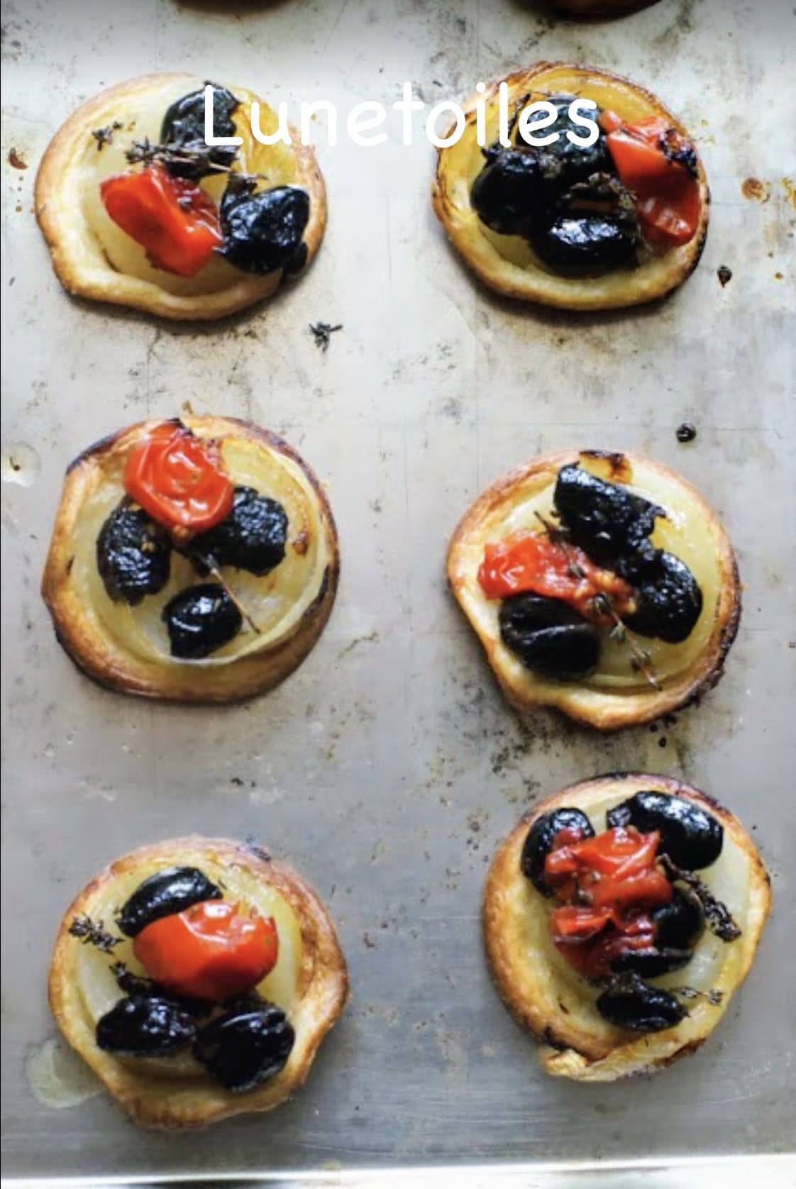 Tartelettes tatin aux oignons caramélisés, olives noires et tomates cerises, cuites au four sur une plaque en métal.