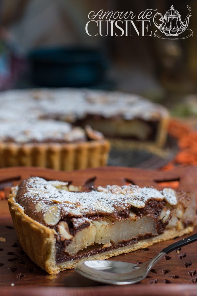 Part de tarte poires et chocolat de Stéphane Glacier, garnie de poires fondantes, crème chocolat onctueuse et amandes effilées, saupoudrée de sucre glace, servie sur un plat en bois
