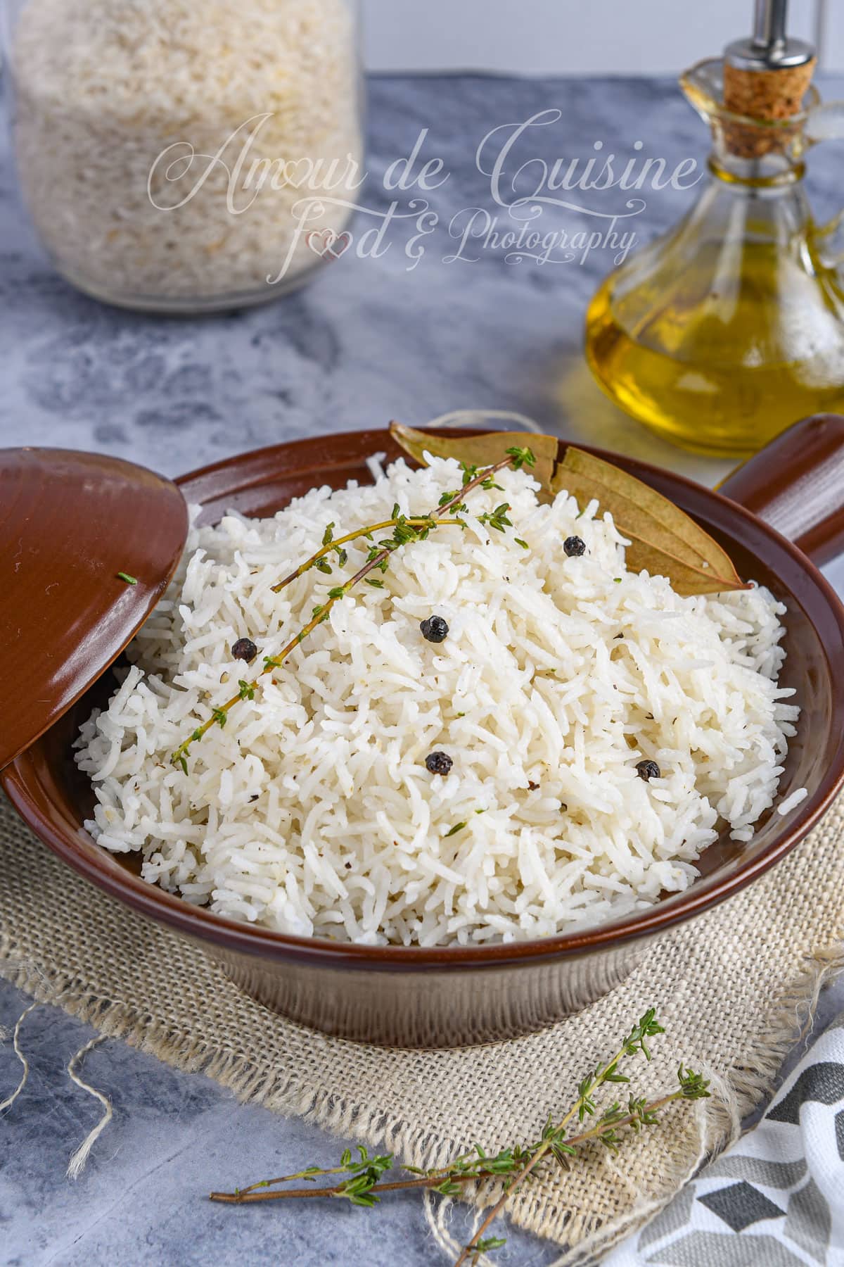 Plat de riz basmati blanc garni de grains de poivre noir, de thym frais et de feuilles de laurier, présenté dans un bol en céramique brune, avec une bouteille d’huile d’olive et un bocal de riz en arrière-plan.