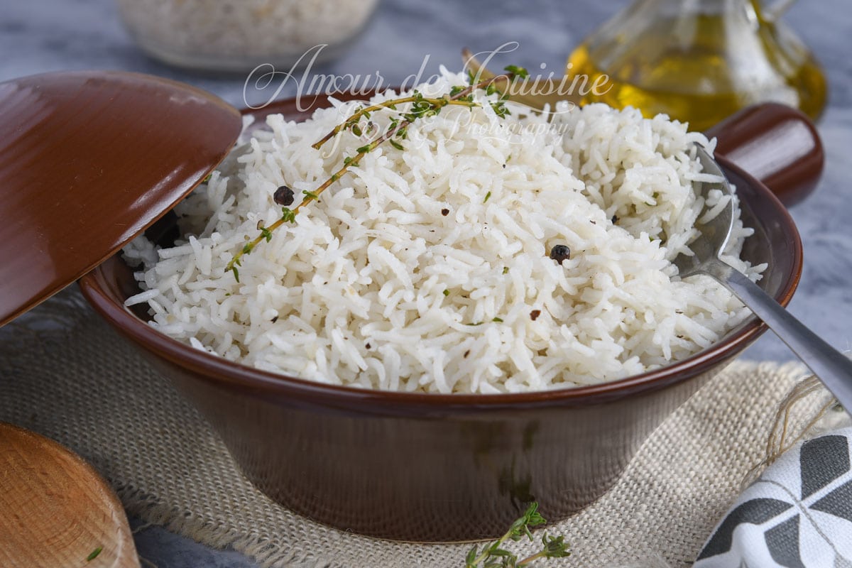 Bol en céramique brune rempli de riz basmati blanc garni de grains de poivre noir et de thym, avec une cuillère en métal et une bouteille d’huile en arrière-plan.