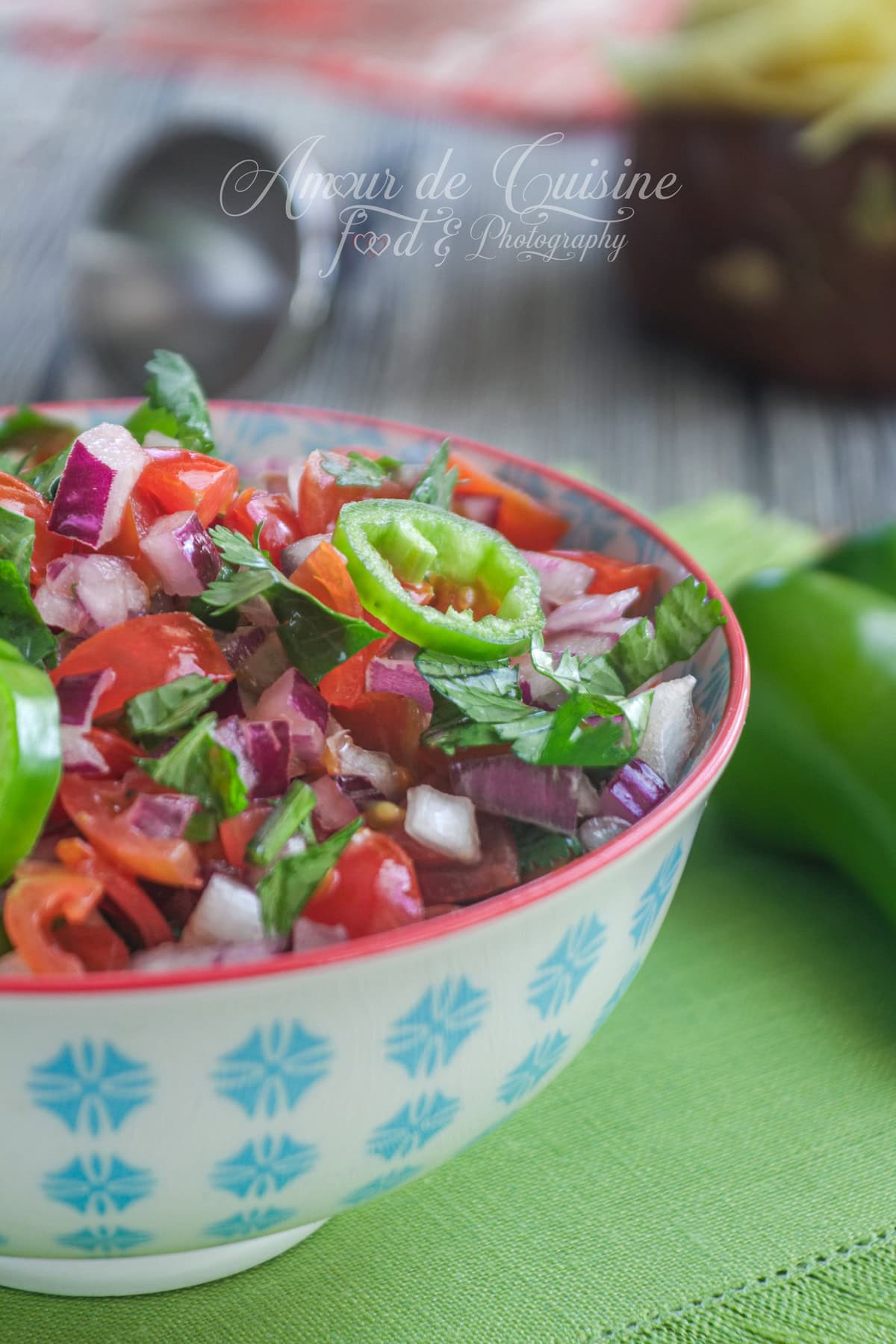 Close-up of a bowl of pico de gallo (fresh salsa) with diced tomatoes, red onion, cilantro and a green chili slice, set on a green napkin with a blurred background (spoon and chips).