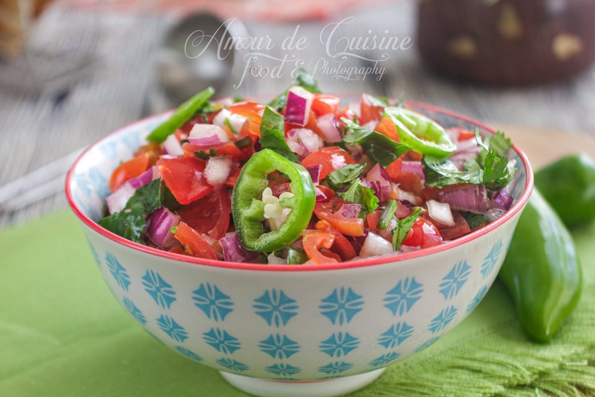 Close-up of a bowl of pico de gallo (fresh salsa) with diced tomatoes, red onion, cilantro and sliced green chili, set on a green cloth with a whole green pepper in the background.