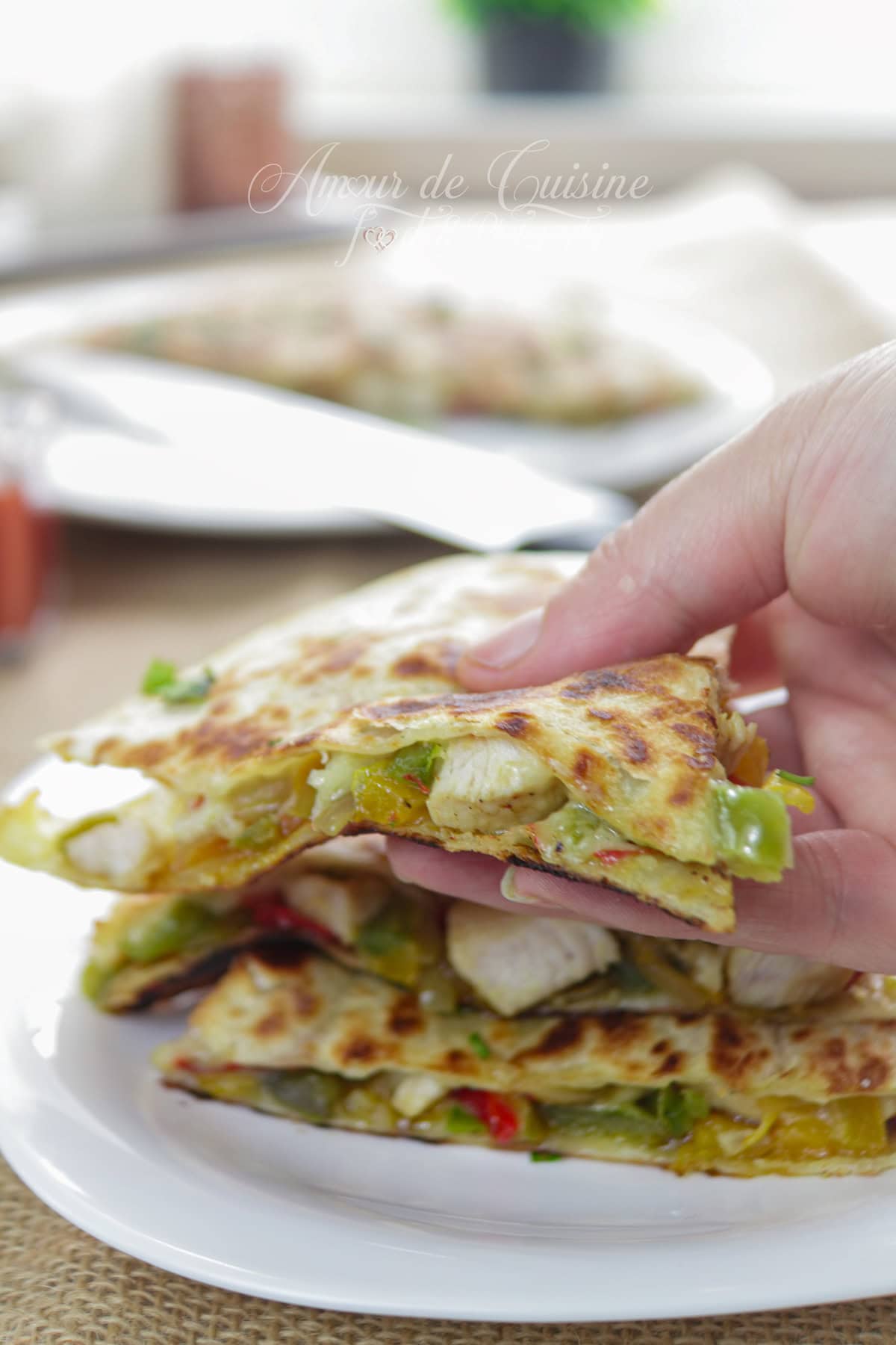 Main qui soulève une part de quesadilla au poulet et poivrons, tortilla dorée et fromage fondant, au-dessus d’une assiette avec d’autres triangles.