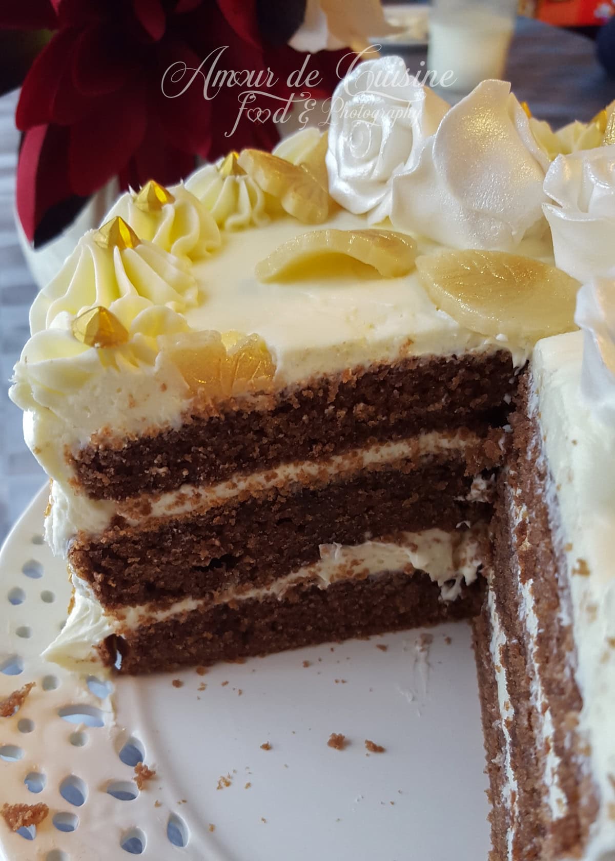 Three-layer chocolate yogurt cake covered in smooth white frosting, decorated with golden sugar leaves and cream rosettes, displayed on a white cake stand.