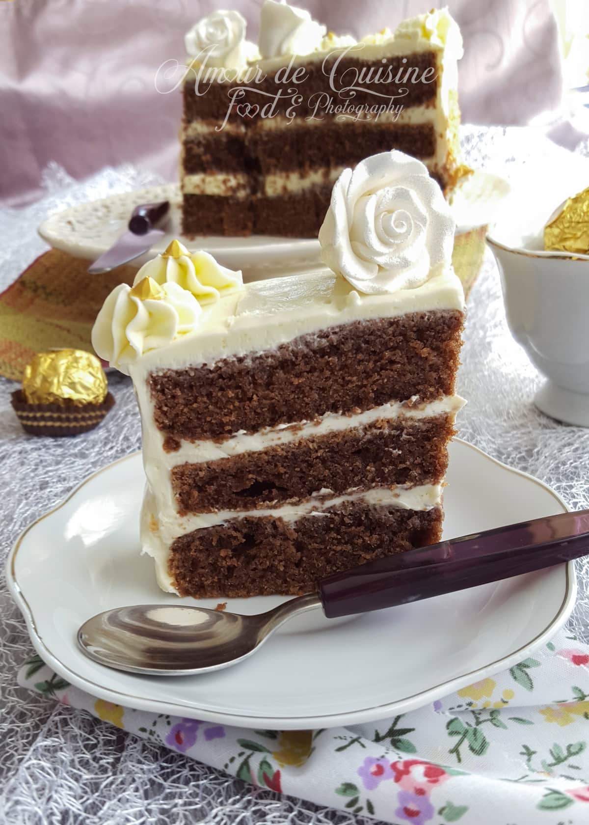 Slice of three-layer chocolate yogurt cake with creamy white frosting, topped with a white rose and golden decorations, served on a plate with a spoon.