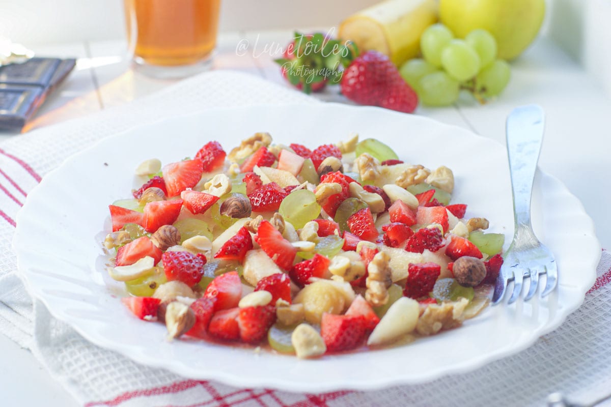 Miam o fruit dans une assiette blanche avec fraises, raisins, banane, pomme et fruits secs pour un petit déjeuner frais.