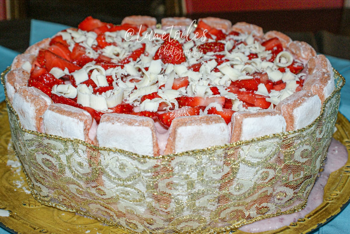 Charlotte aux fraises garnie de biscuits roses de Reims, surmontée de fraises fraîches et copeaux de chocolat blanc, entourée d’un ruban décoratif doré et présentée sur un plateau doré