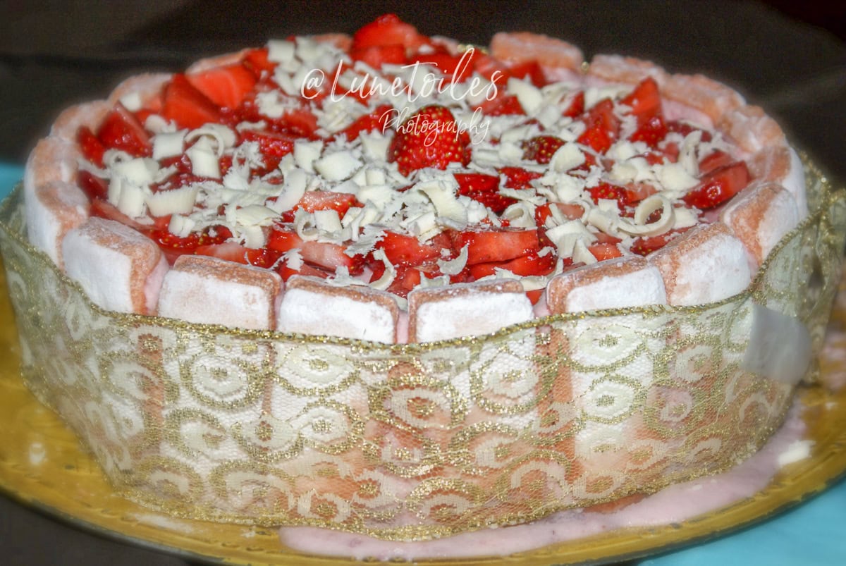 Gâteau charlotte aux fraises décoré de fraises fraîches et copeaux de chocolat blanc, entouré de biscuits roses de Reims et d’un ruban doré à motifs, présenté sur un plat doré avec un nappage coulant légèrement sur les bords