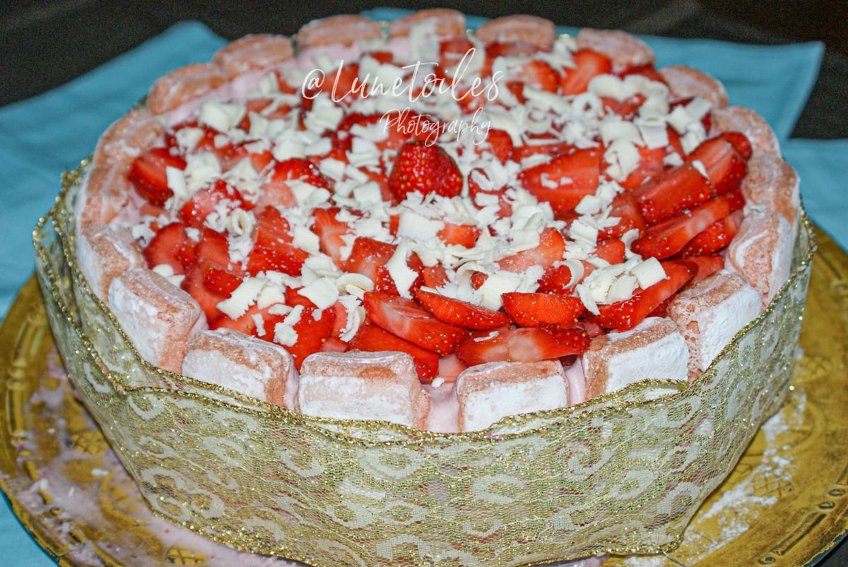 Gâteau charlotte aux fraises décoré de fraises fraîches et copeaux de chocolat blanc, entouré de biscuits roses de Reims et d’un ruban doré à motifs, présenté sur un plat doré avec un nappage coulant légèrement sur les bords